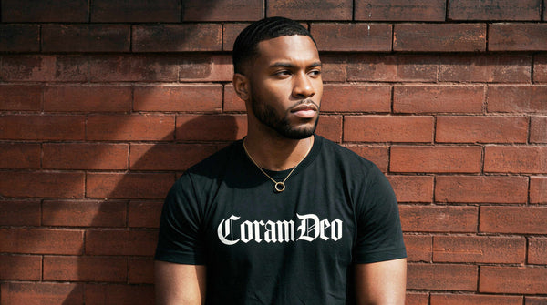 Man wearing a black t-shirt with 'Coram Deo' text against a brick wall.